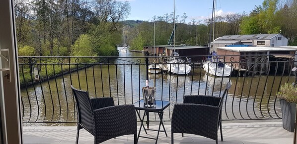 A balcony with wicker chairs and a table, overlooking a river with boats and a dock. - Modern Waterfront Boathouse With Beautiful Balcony On Windermere Marina (Bowness-on-Windermere)