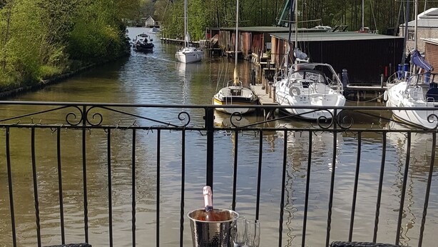 Un balcon agrémenté de chaises et d’une table en osier, donnant sur une rivière paisible où des bateaux sont amarrés le long de la rive.