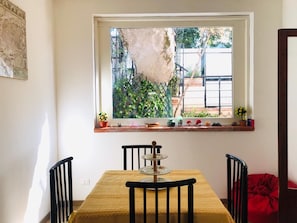 A dining area with a table and chairs, a window with a view of greenery, and a shelf with various items.