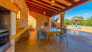 Une terrasse extérieure avec un mur en pierre, des poutres en bois, une longue table avec des chaises et un espace piscine avec des chaises longues.