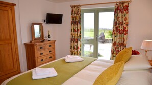 A hotel room with a bed, a wooden dresser, a television on the wall, and a view of the outdoors through a door. - Hazel Cottage Purpose built wheelchair friendly cottage (Truro)