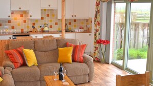 A modern living room with a sofa, coffee table, and a dining area with a table and chairs. A kitchen with white cabinets and a tiled backsplash is visible in the background. - Hazel Cottage Purpose built wheelchair friendly cottage (Truro)