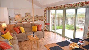 A living room with a sofa, a wooden coffee table, and a dining table with a bowl of fruit and croissants. - Hazel Cottage Purpose built wheelchair friendly cottage (Truro)