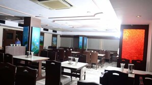 A restaurant with multiple dining tables and booths, a counter with a person behind it, and a large abstract red wall. - Jaya Residency (Kakinada)