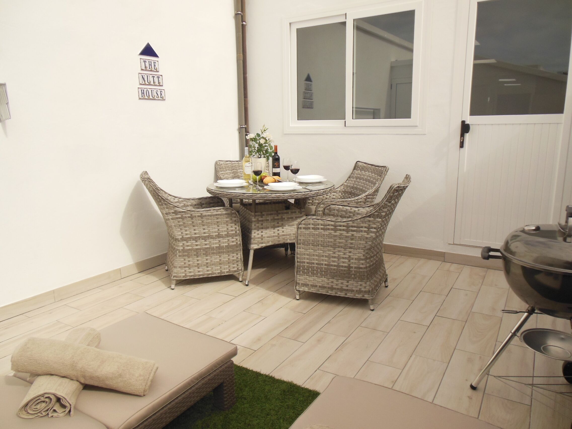 Un coin repas avec des chaises en osier et une table dressée pour quatre personnes, un petit parterre avec une serviette enroulée, et un panneau accroché au mur.