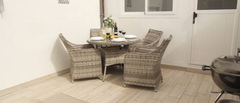 Un coin repas avec des chaises en osier et une table dressée pour quatre personnes, un petit parterre avec une serviette enroulée, et un panneau accroché au mur.