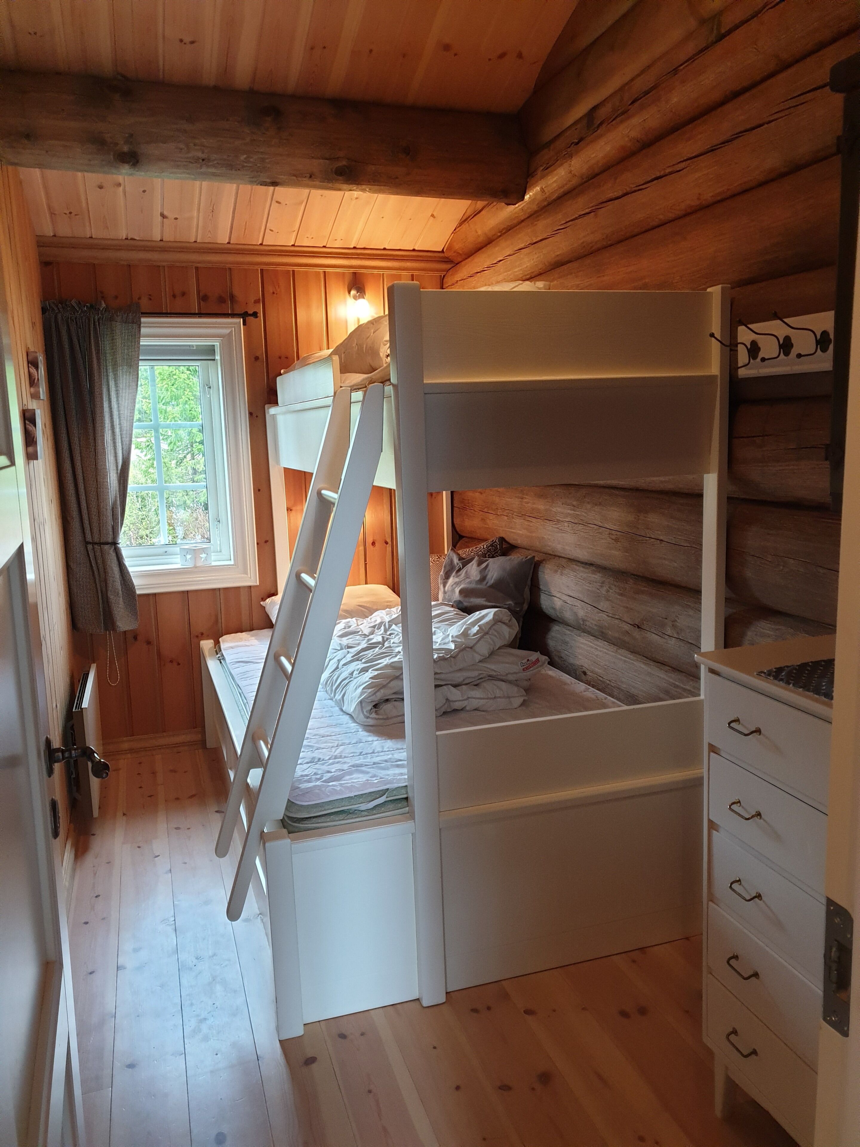 A bunk bed in a wooden cabin room with a window, a ladder, and a dresser.