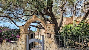 Una puerta de piedra con un arco de metal que da a un sendero pavimentado, rodeado de abundante vegetación y un muro de piedra.