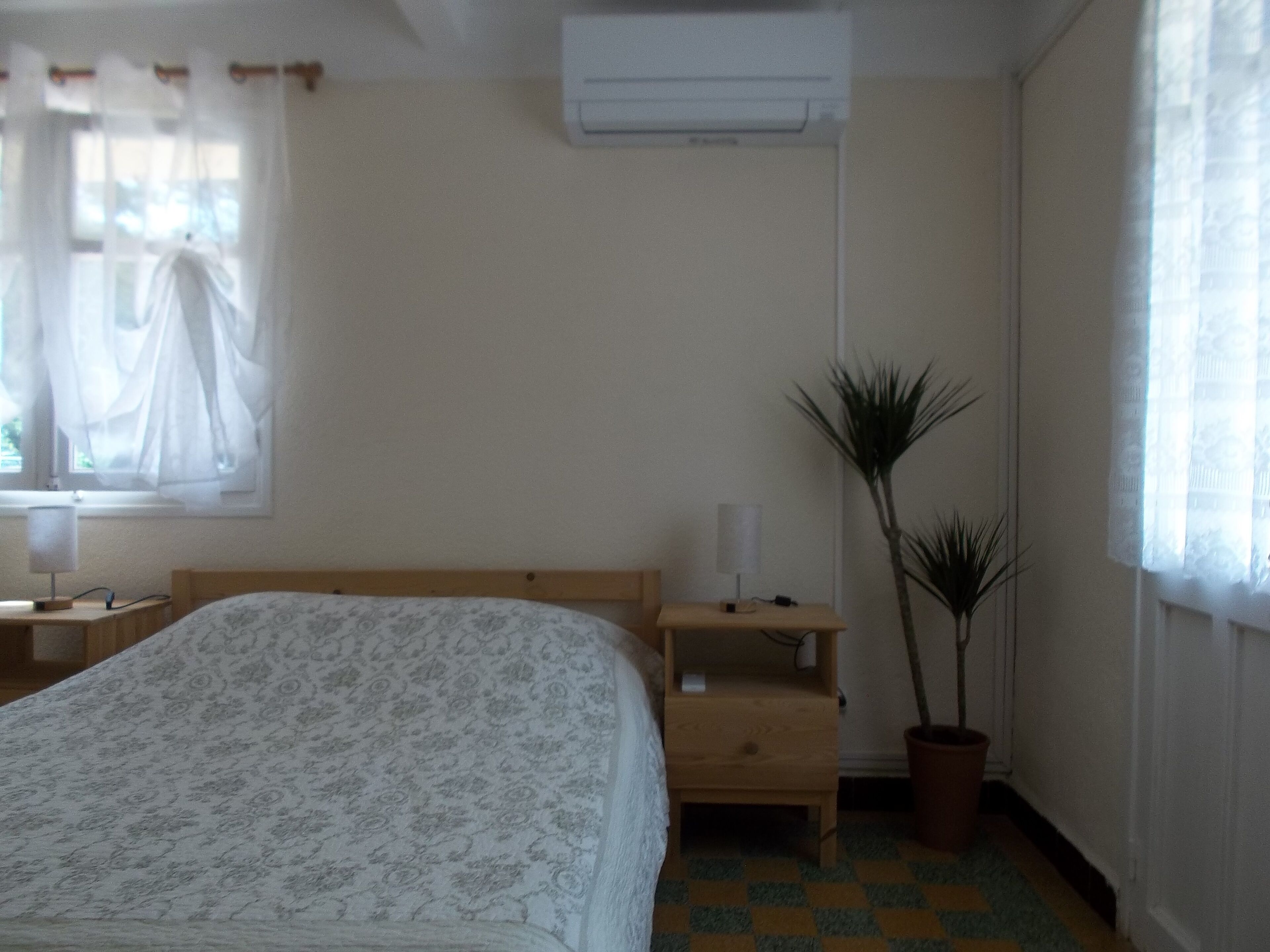 A bedroom with a bed, a bedside table, a nightstand, a potted plant, and a window with curtains.