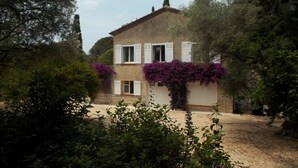 Una casa de dos pisos con contraventanas blancas, rodeada de abundante vegetación y árboles.