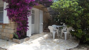 Un patio con sillas de plástico blanco y una mesa, rodeado de piso de piedra y abundante vegetación.
