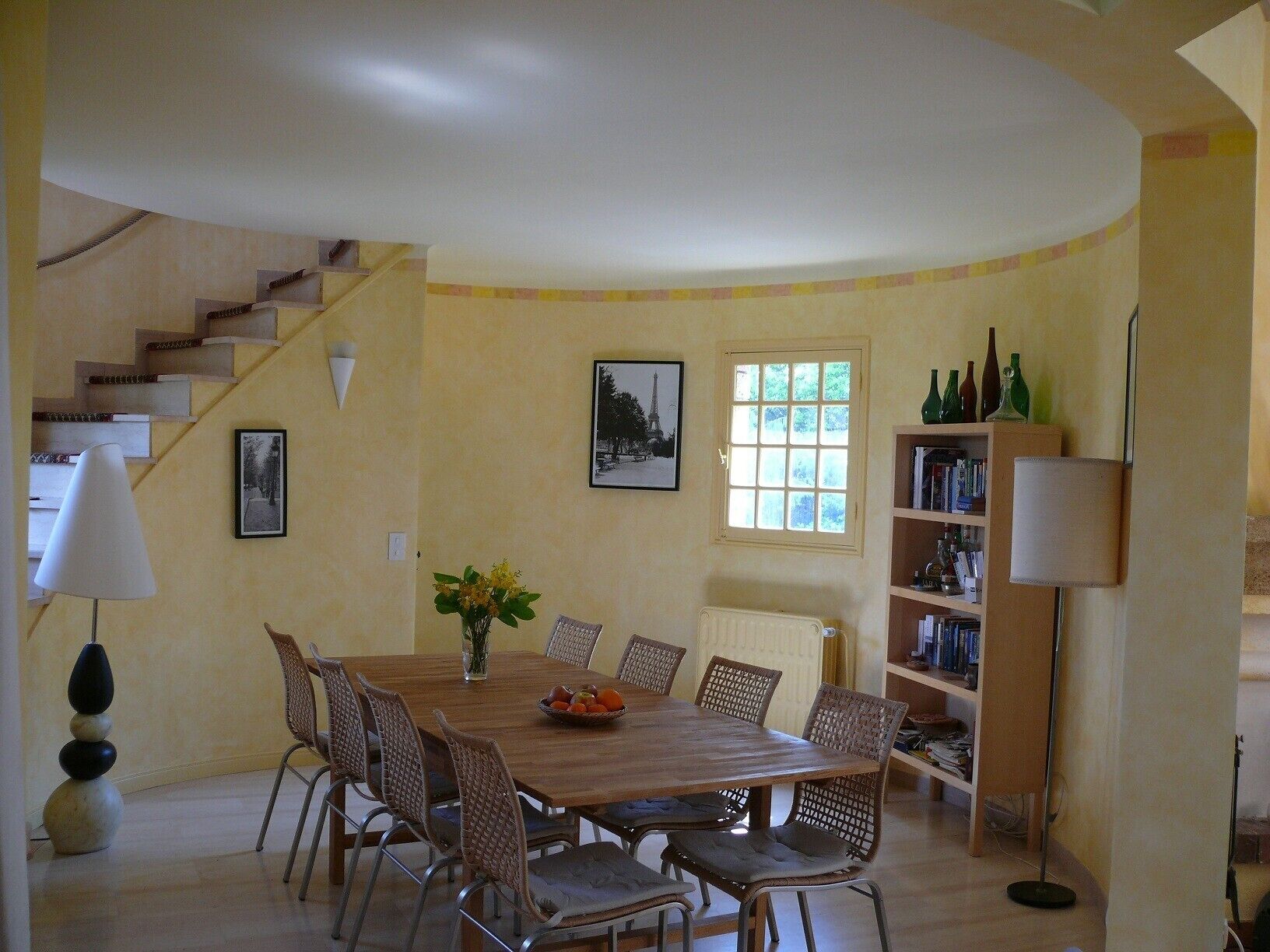 Un comedor con mesa de madera y sillas de mimbre, una escalera, una estantería y un radiador.