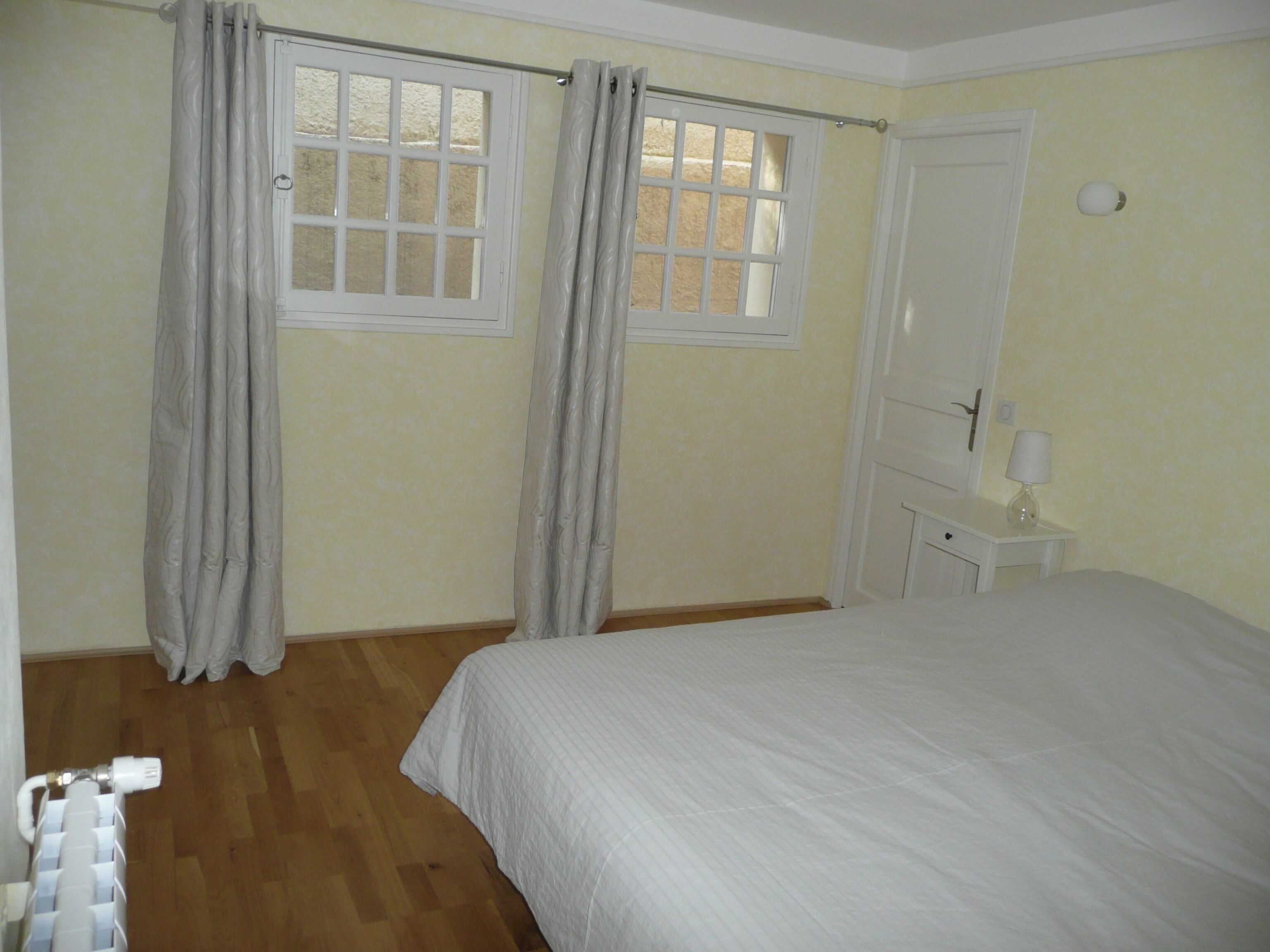 A bedroom with a bed, two windows with curtains, a radiator, and a nightstand with a lamp.