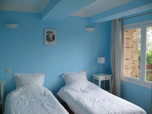 A room with two single beds, a small bedside table with a lamp, a framed picture on the wall, and a window with a view of a brick wall.
