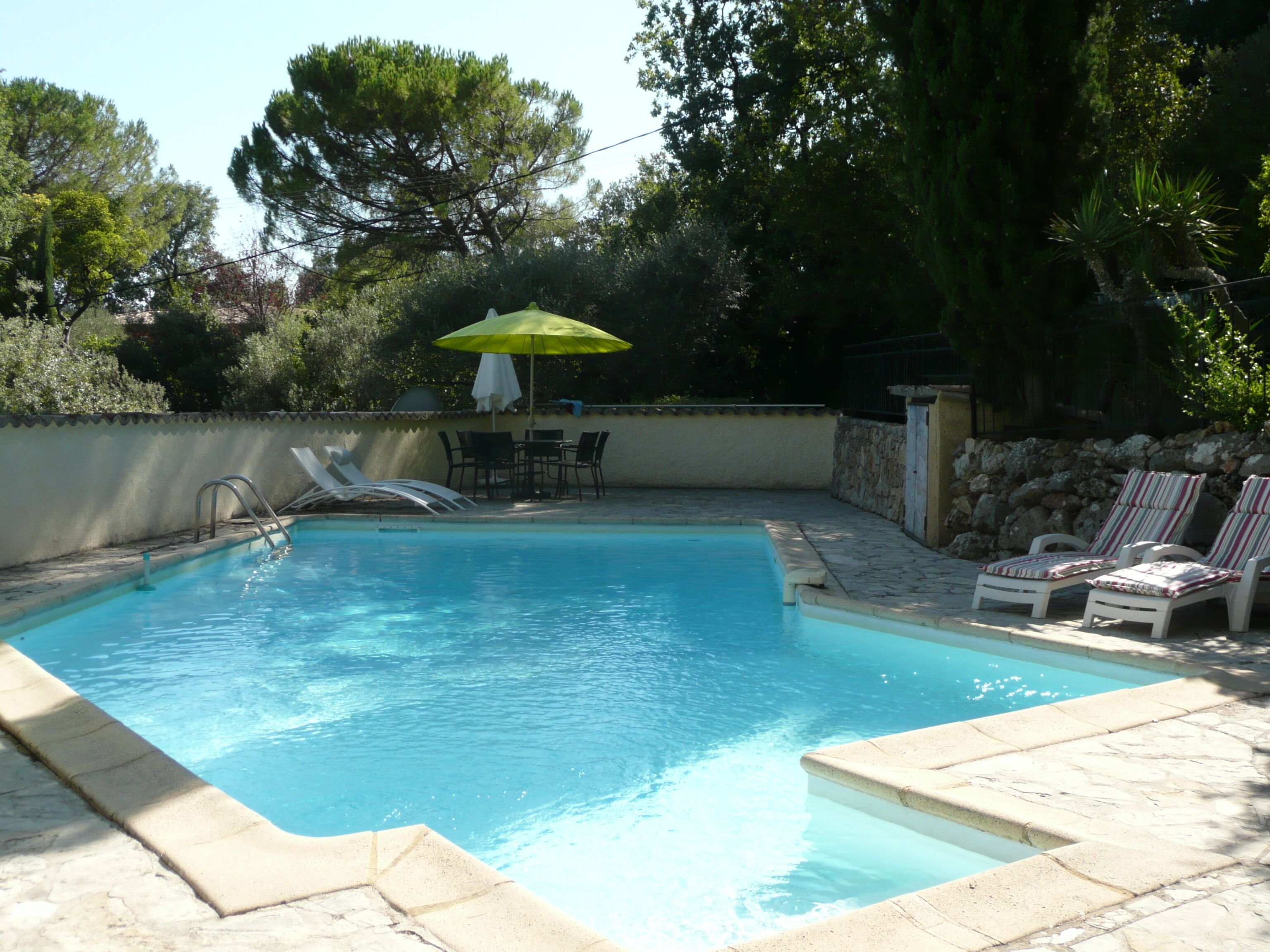 Una piscina rectangular con borde de piedra, rodeada de sillones de descanso y una mesa con sombrilla.