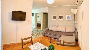 A living room with a sofa, a TV mounted on the wall, a dining table with chairs, and a lamp.