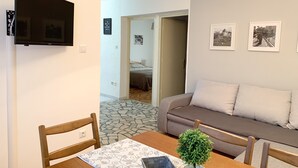 A living room with a sofa, a dining table with chairs, a TV mounted on the wall, and a small potted plant.