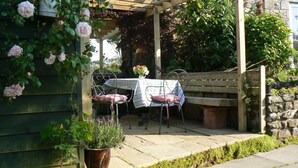 A covered patio with a wooden table and chairs, surrounded by greenery and a stone wall. - Romantic Retreat (Penryn)