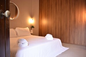 Een net opgemaakt bed met witte lakens en handdoeken, een nachtkastje met een plant en een houten paneelwand.