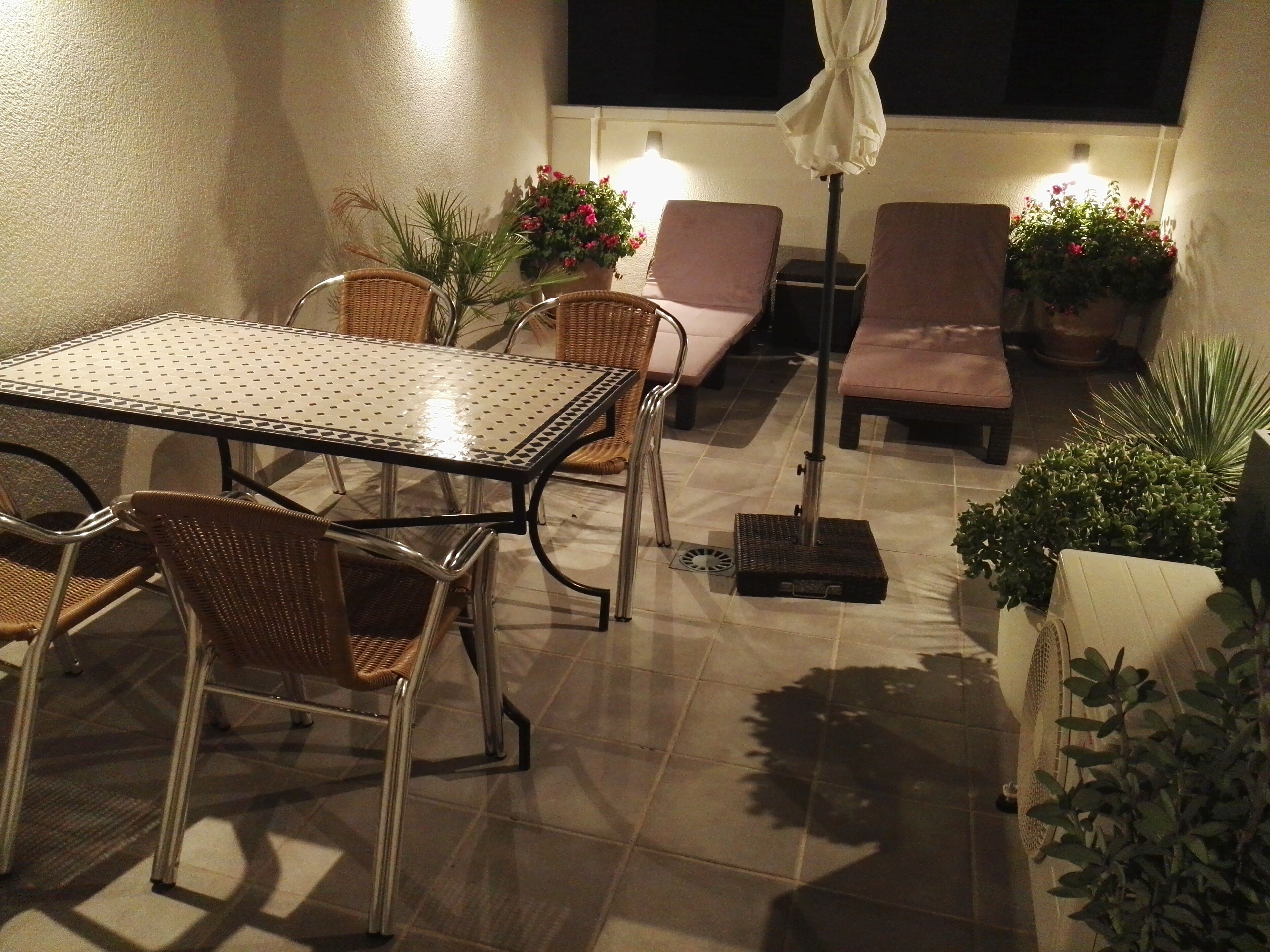 An outdoor patio with a metal table and wicker chairs, surrounded by potted plants and a floor lamp.