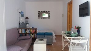Una habitación pequeña y bien iluminada con sofá, estantería, mesita, cama, ventana con marco decorativo y televisor montado en la pared.
