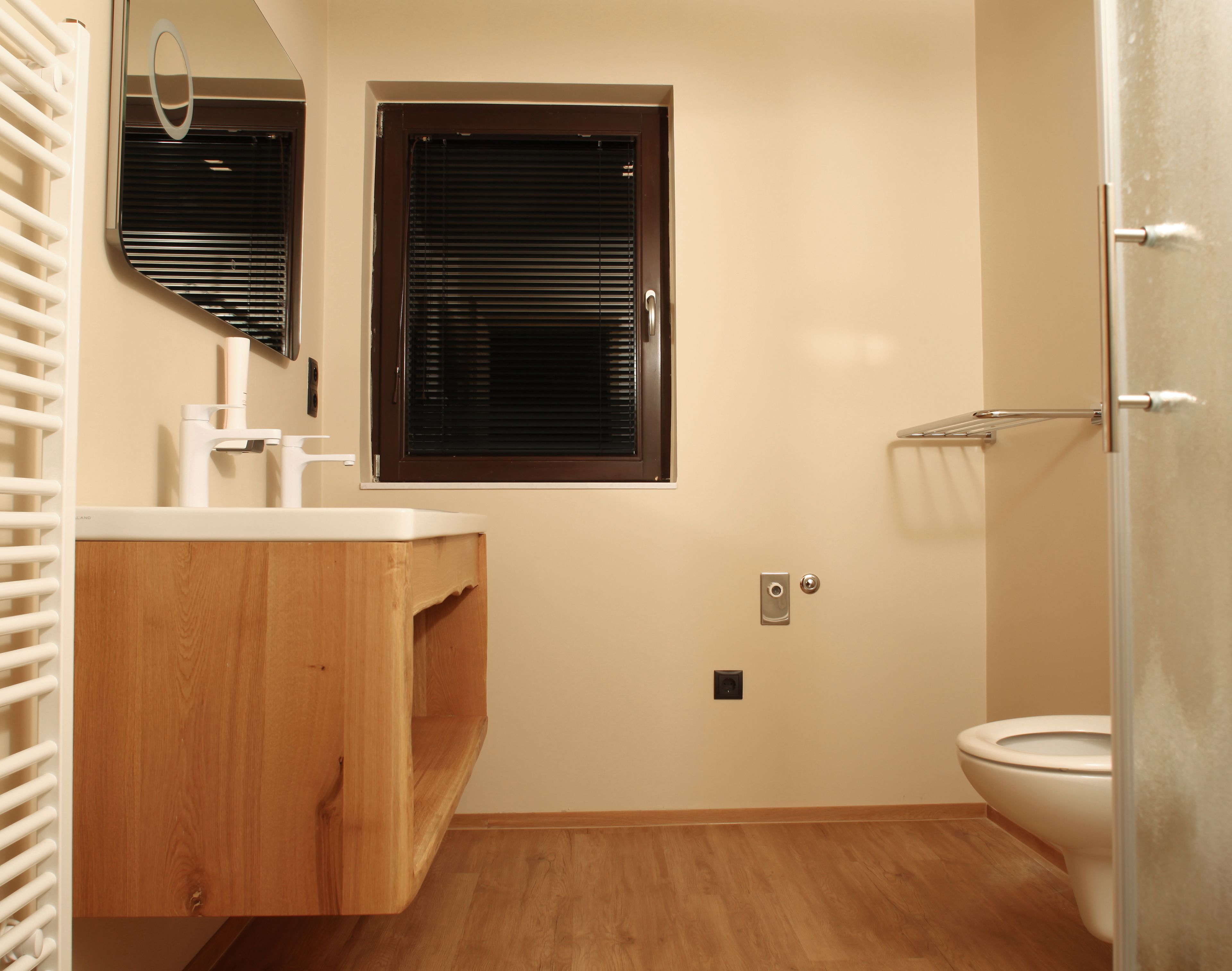 A bathroom with a wooden vanity, a window with blinds, a toilet, and a shower area.