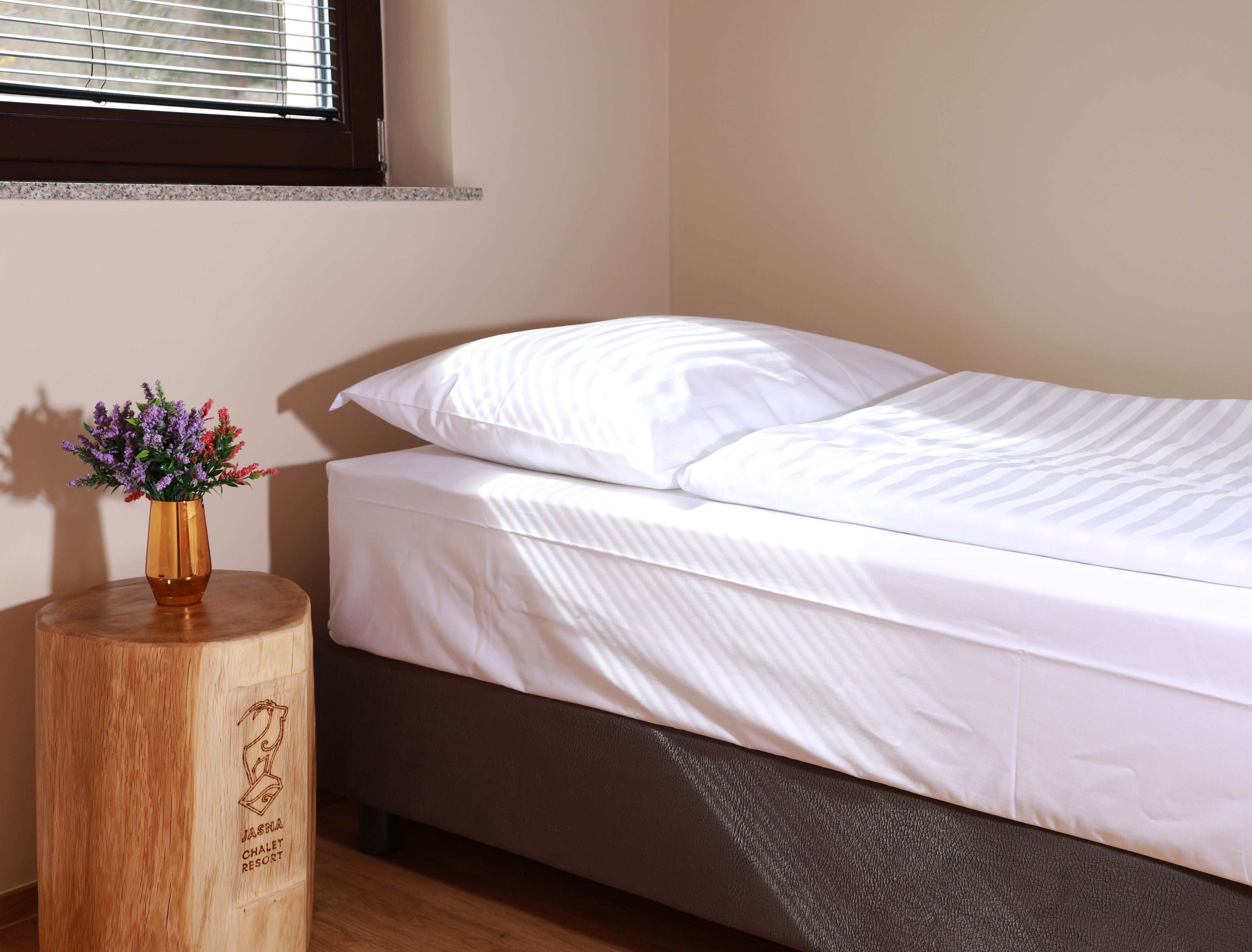 Uma cama com roupa de cama branca, uma mesa de cabeceira de madeira com um vaso de flores e uma janela com persianas.