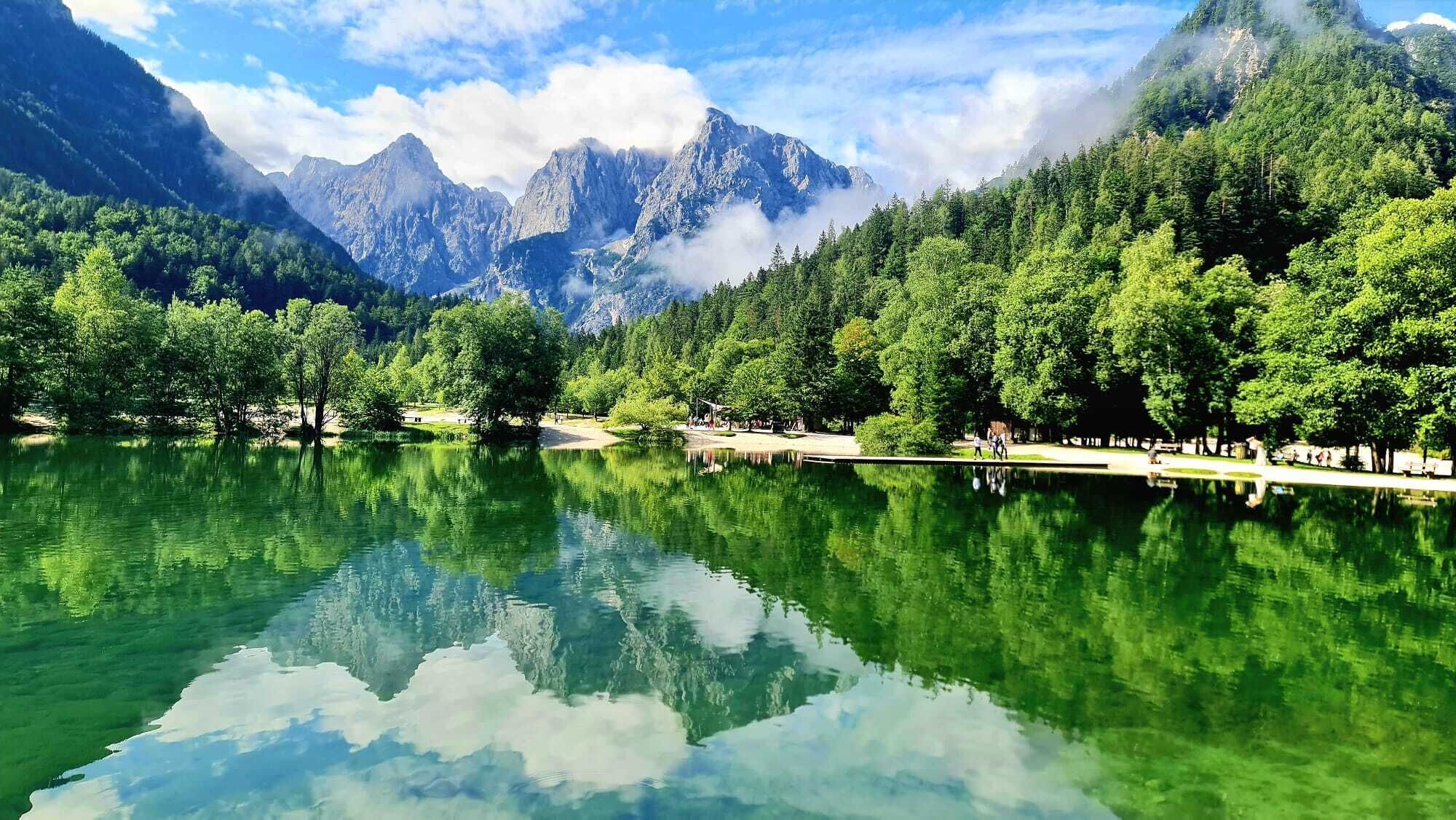 A serene lake with clear reflections, surrounded by lush green trees and mountains in the background.