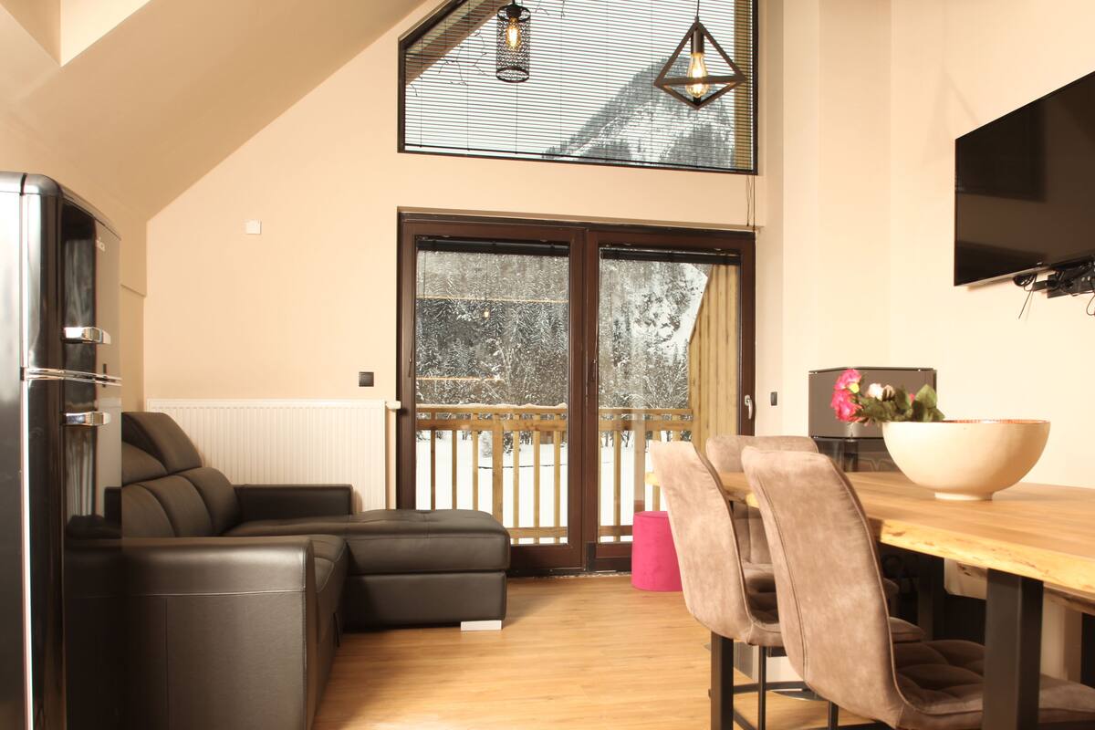 A modern kitchen with a dining area, a large window with a view of snow-covered trees, and a balcony with a wooden railing.