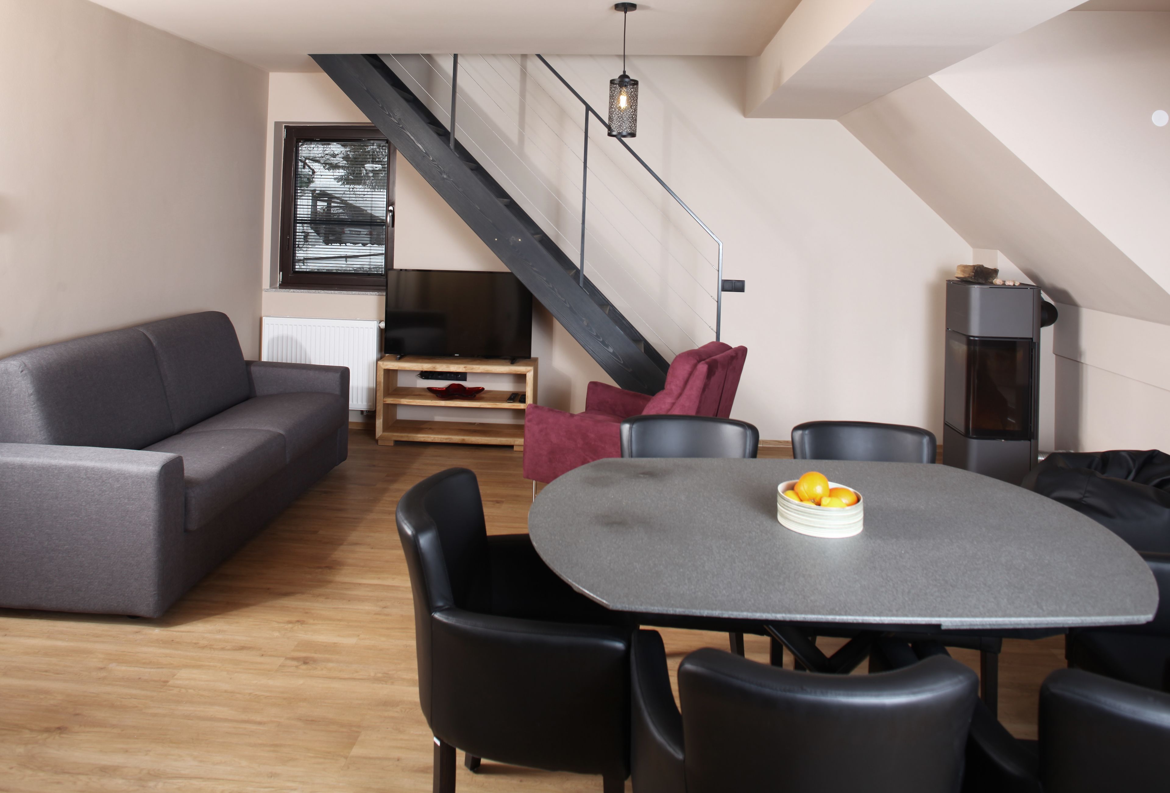 A modern living room with a gray sofa, a dining table with chairs, a staircase, and a TV.
