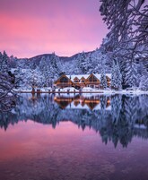 Una cabaña con varias cabañas de madera rodeada de árboles nevados y un lago congelado que refleja los edificios y el cielo.