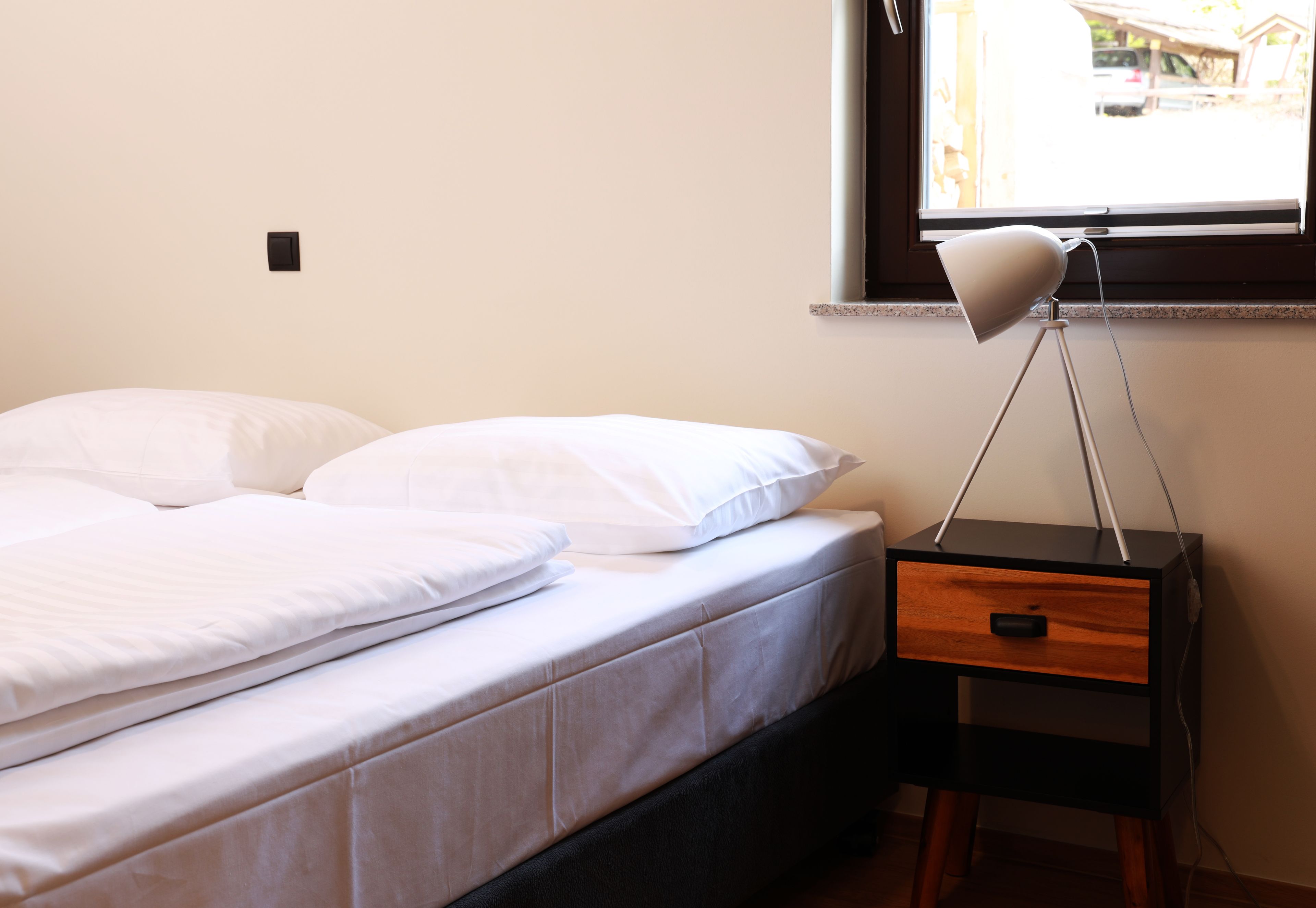 A neatly made bed with white linens, a bedside table with a lamp, and a window with a view of the outside.