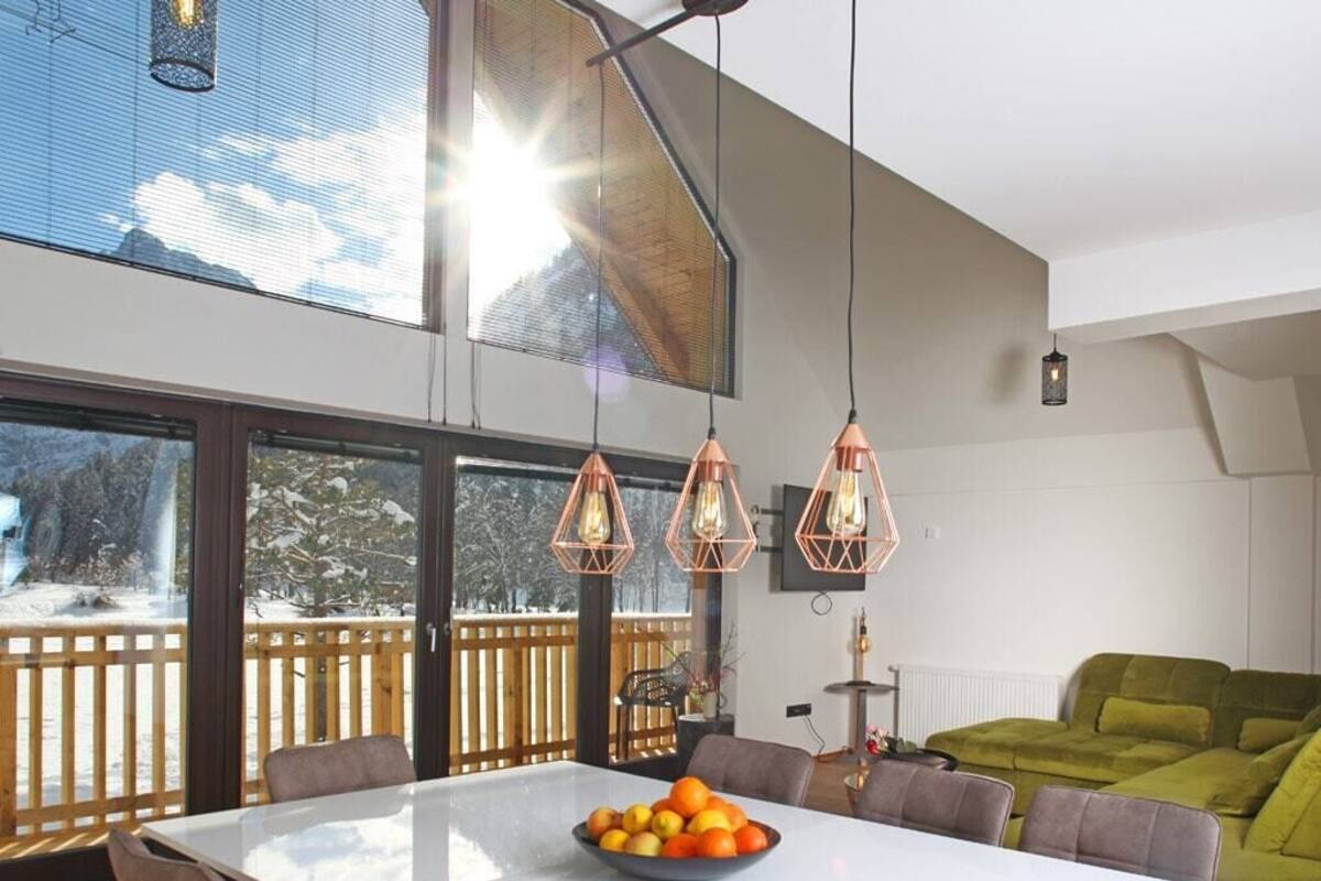 A modern kitchen with a large dining table, pendant lights, and a view of a snowy landscape through large windows.