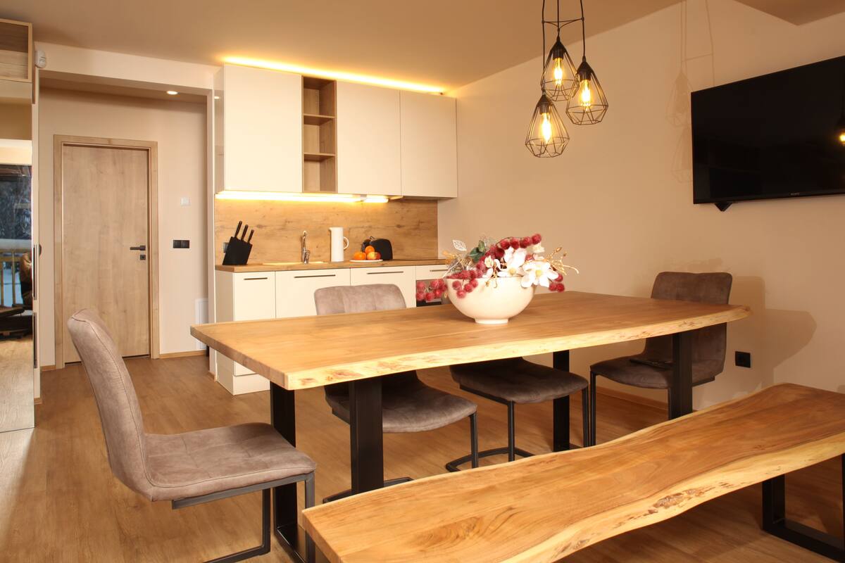 A modern kitchen with white cabinets, a wooden dining table, and a TV mounted on the wall.