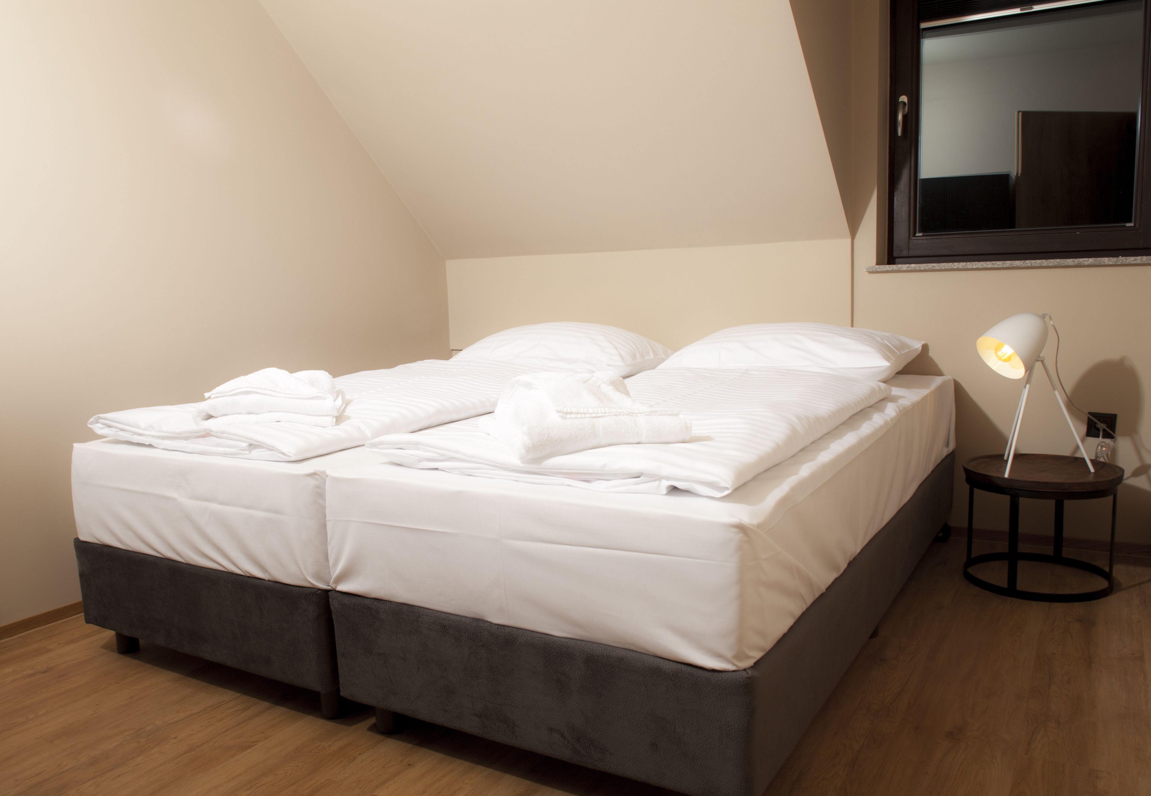 A neatly made bed with white linens and a bedside lamp, in a room with wooden flooring and a sloped ceiling.