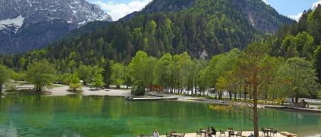 A lakeside terrace with outdoor seating, surrounded by forested mountains and a clear sky.