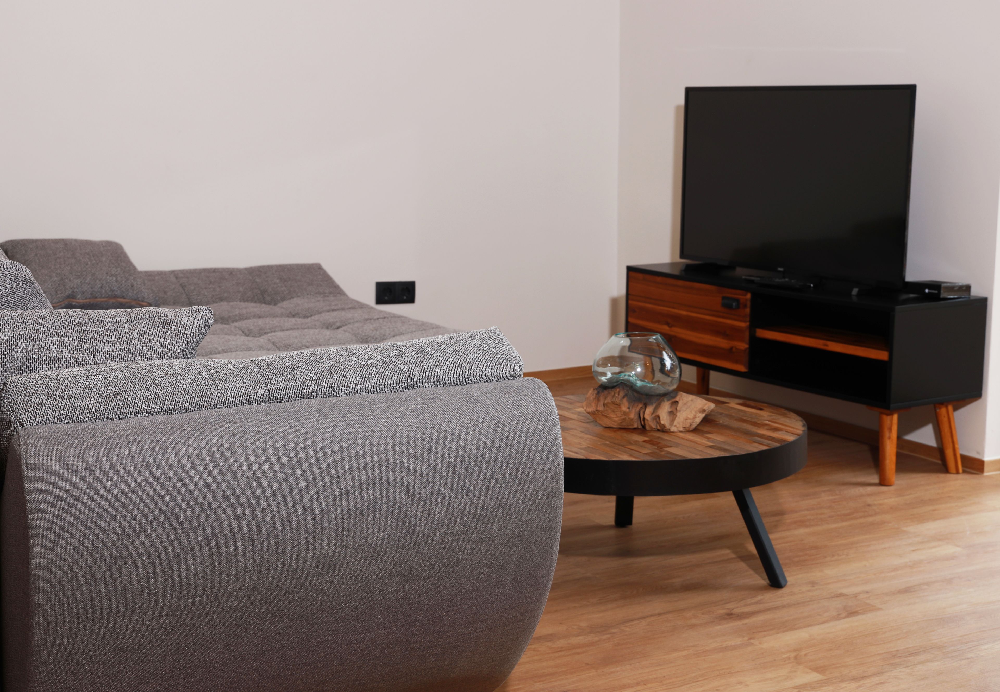 Una sala moderna con un sofá gris, una mesa de centro redonda, un televisor de pantalla plana sobre un mueble de madera y una pequeña mesita de madera con un jarrón de cristal.