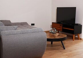 Una sala moderna con un sofá gris, una mesa de centro redonda, un televisor de pantalla plana sobre un mueble de madera y una pequeña mesita de madera con un jarrón de cristal.