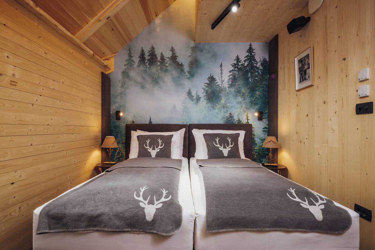 A wooden cabin bedroom with two beds, a forest mural on the wall, and a mountain view.