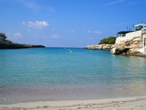 A coastal scene with clear water, a sandy beach, and rocky cliffs.