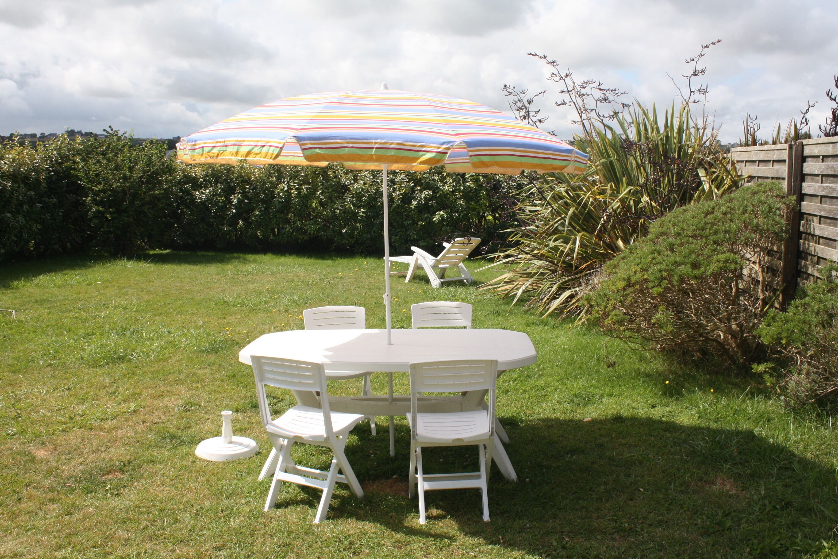 Un ensemble de mobilier de jardin blanc, comprenant une table et des chaises, agrémenté d’un parasol rayé, le tout entouré de verdure et d’une clôture en bois.