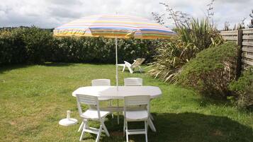Un ensemble de mobilier de jardin blanc, comprenant une table et des chaises, agrémenté d’un parasol rayé, le tout entouré de verdure et d’une clôture en bois.