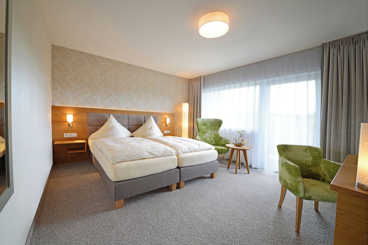 Ein Hotelzimmer mit einem Doppelbett, zwei Nachttischen, einem grünen Sessel, einem kleinen runden Tisch und einem großen Fenster mit Vorhängen.