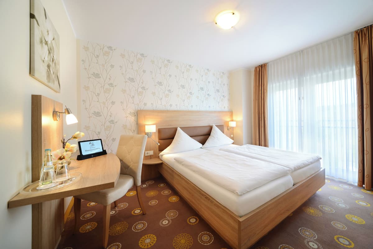 Ein Hotelzimmer mit einem großen Bett, einem Schreibtisch mit Computer, einem Sessel und einem kleinen Tisch mit Blumenvase.
