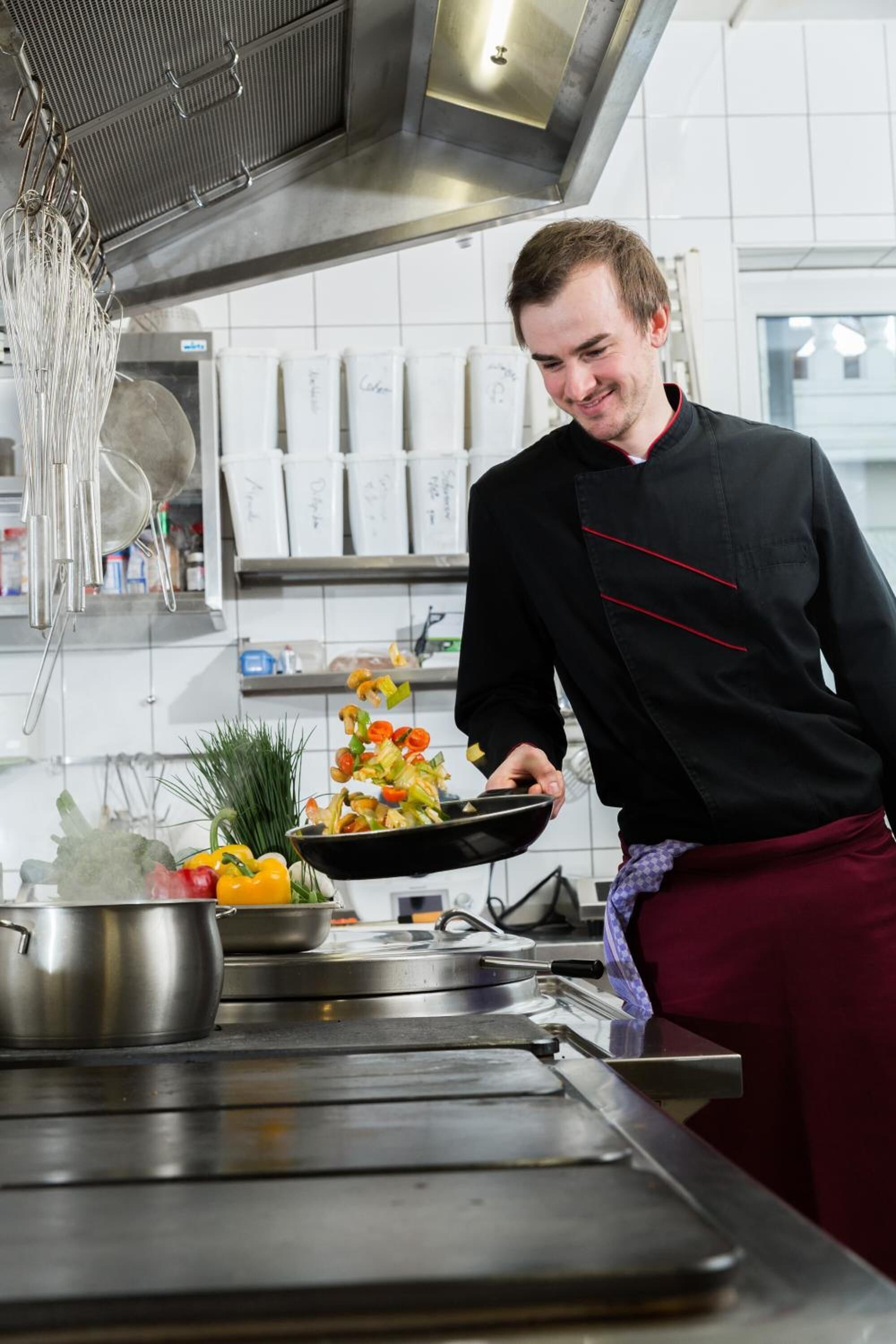 Un chef en una cocina profesional con superficies de acero inoxidable, una olla sobre la estufa y diversos utensilios de cocina.