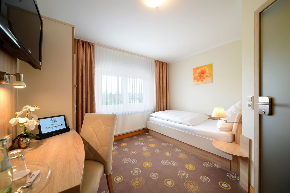 Ein Hotelzimmer mit einem Bett, einem Schreibtisch mit Computer, einem Sessel, einem Fernseher und einem Fenster mit Vorhängen.