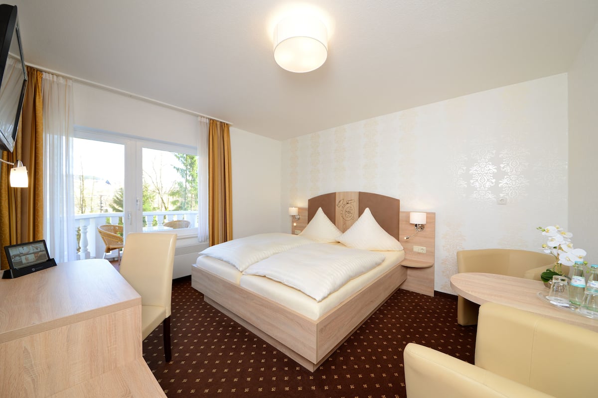 Ein Hotelzimmer mit einem Bett, einem Schreibtisch, einem Sessel, einem kleinen Tisch mit Blumen und einem Fenster mit Vorhängen.