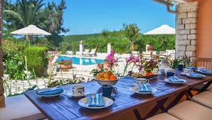 Mesa dispuesta con platos, tazas y un bol de fruta, con vista a una piscina y un jardín exuberante.