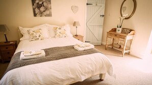 A neatly made bed with a quilt, pillows, and a folded towel. There is a bedside table with a lamp, a dresser with a mirror, and a door with a heart-shaped decoration.