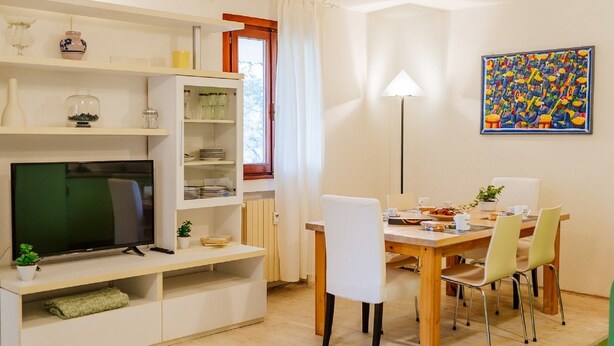 A modern living room with a flat-screen TV, a dining table with chairs, a bookshelf, and a painting on the wall. - Welc-om Green Cottage (Teolo)